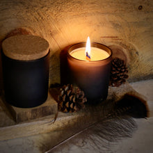 Load image into Gallery viewer, beeswax candle in frosted black  glass jar with cork lid. A lite candle burns on a marble counter with pinecones and turkey feathers in photo display